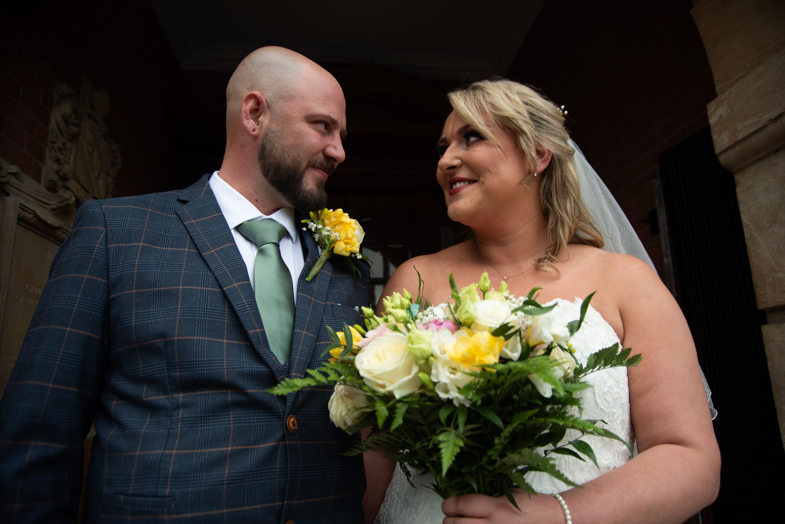 Bride in white dress holds a bouquet while sharing a moment with groom in checkered suit.