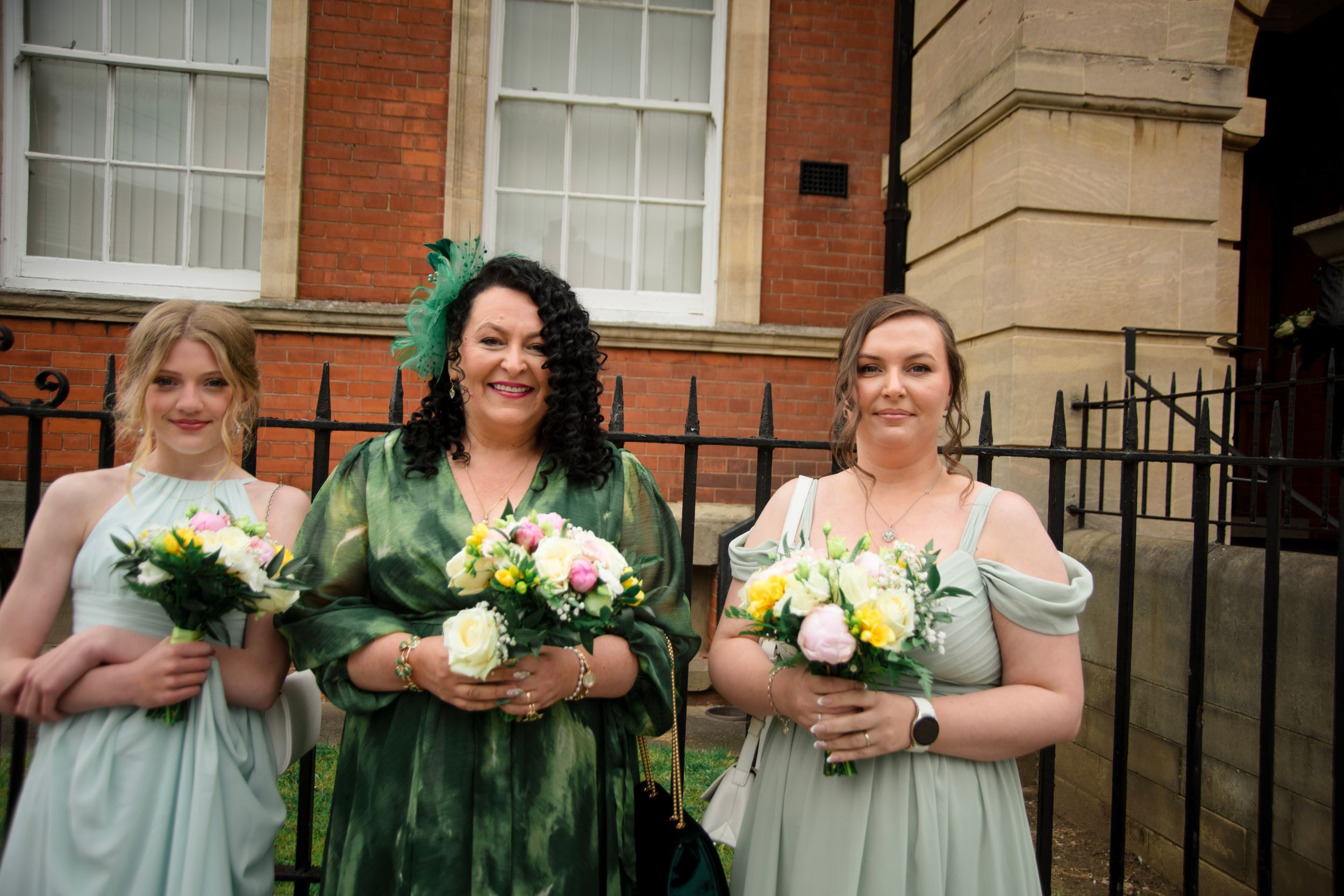 Three bridesmaids in mint green dresses stand together holding white and yellow bouquets outside a brick building.