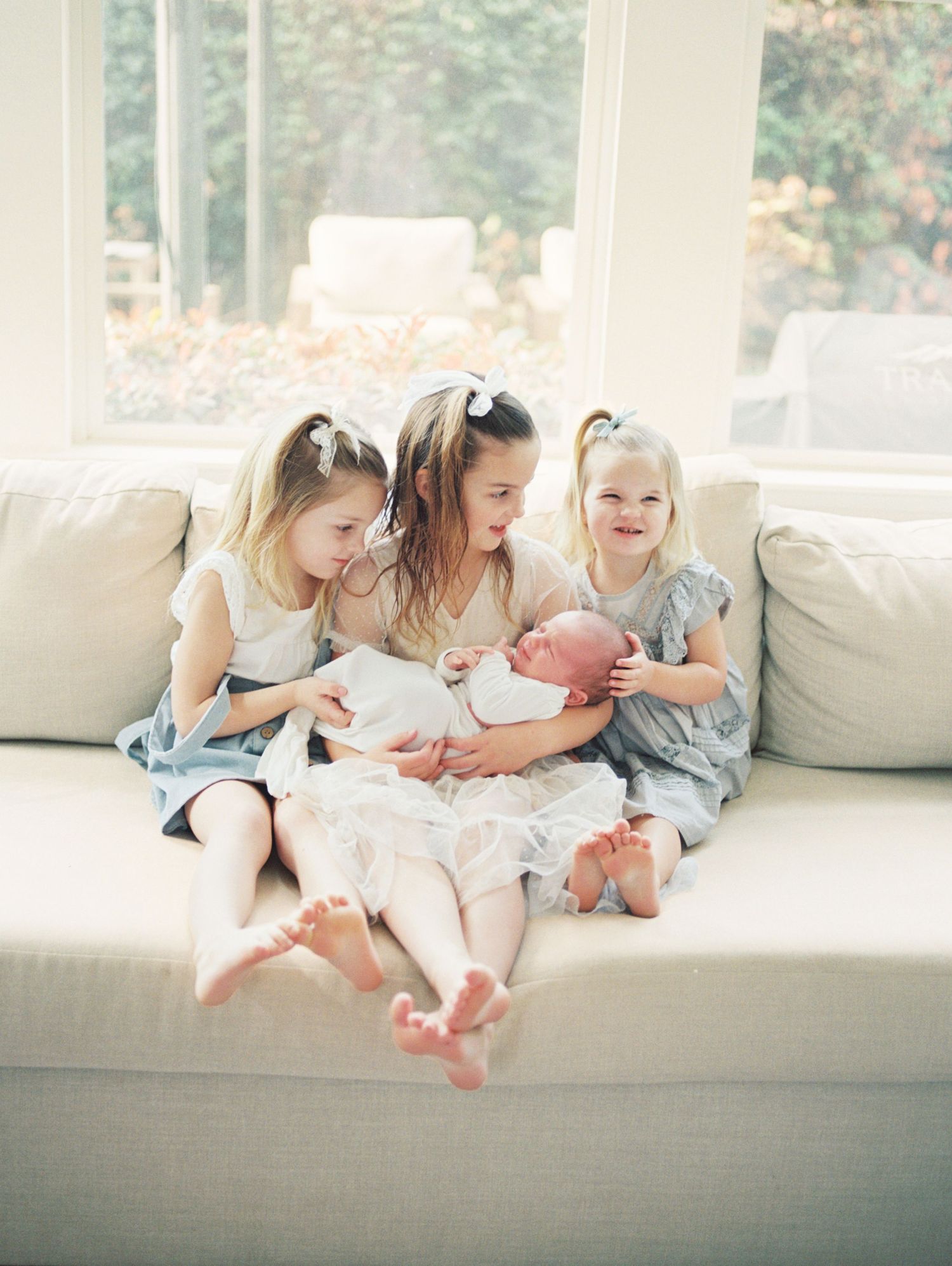 Three siblings sit together on a white couch by a window, sharing a sweet moment while looking down at something.