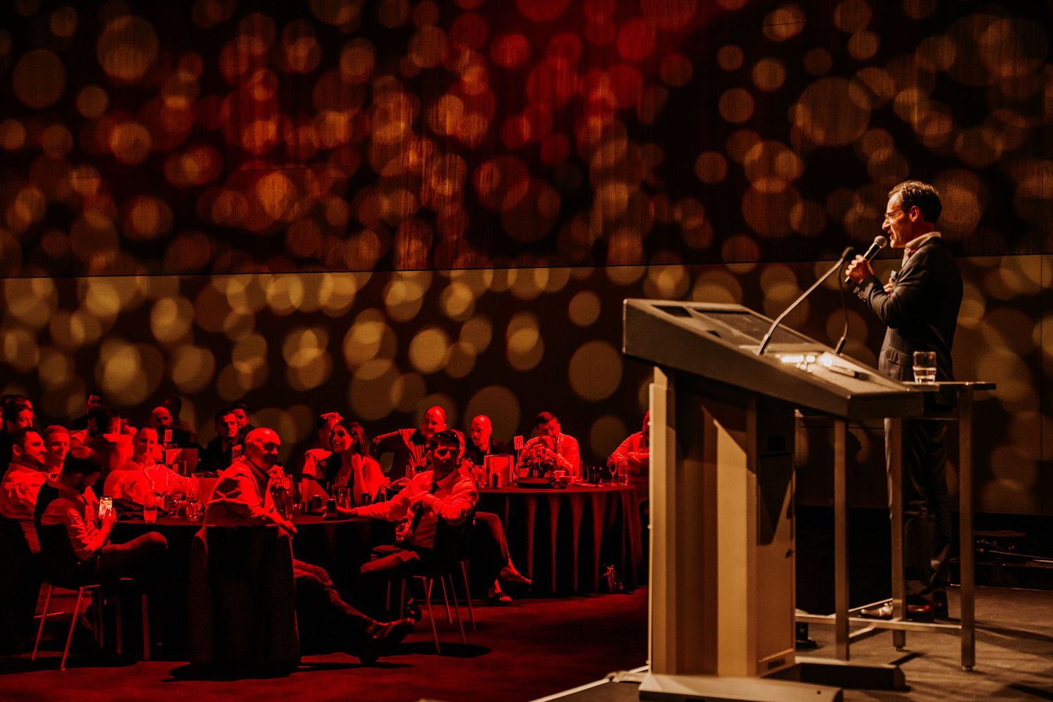 A speaker stands at a podium on stage with dramatic red lighting and a blurred audience in the background.