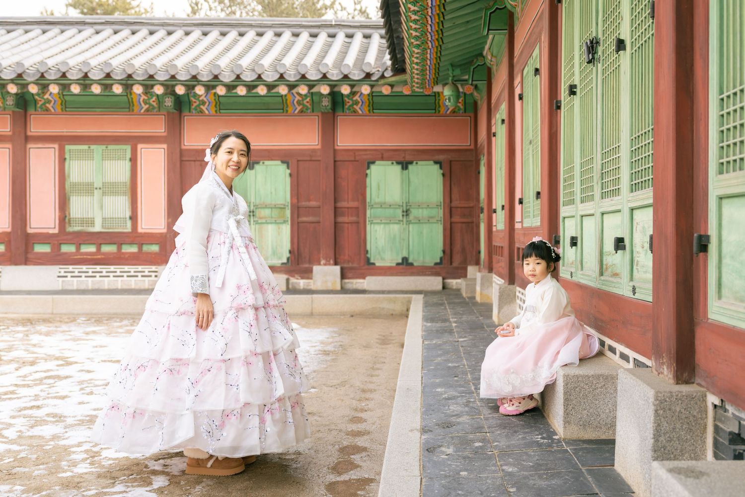 Two people wearing traditional Korean hanbok dresses pose at a historic palace courtyard with traditional architecture.