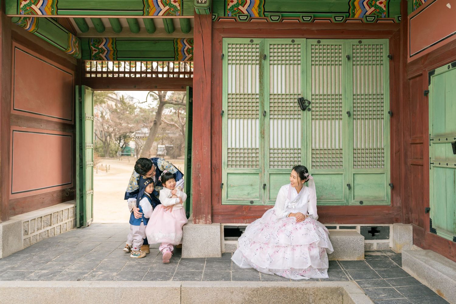 People wearing traditional Korean hanbok clothing sit in a traditional palace doorway with mint green doors and pink walls.