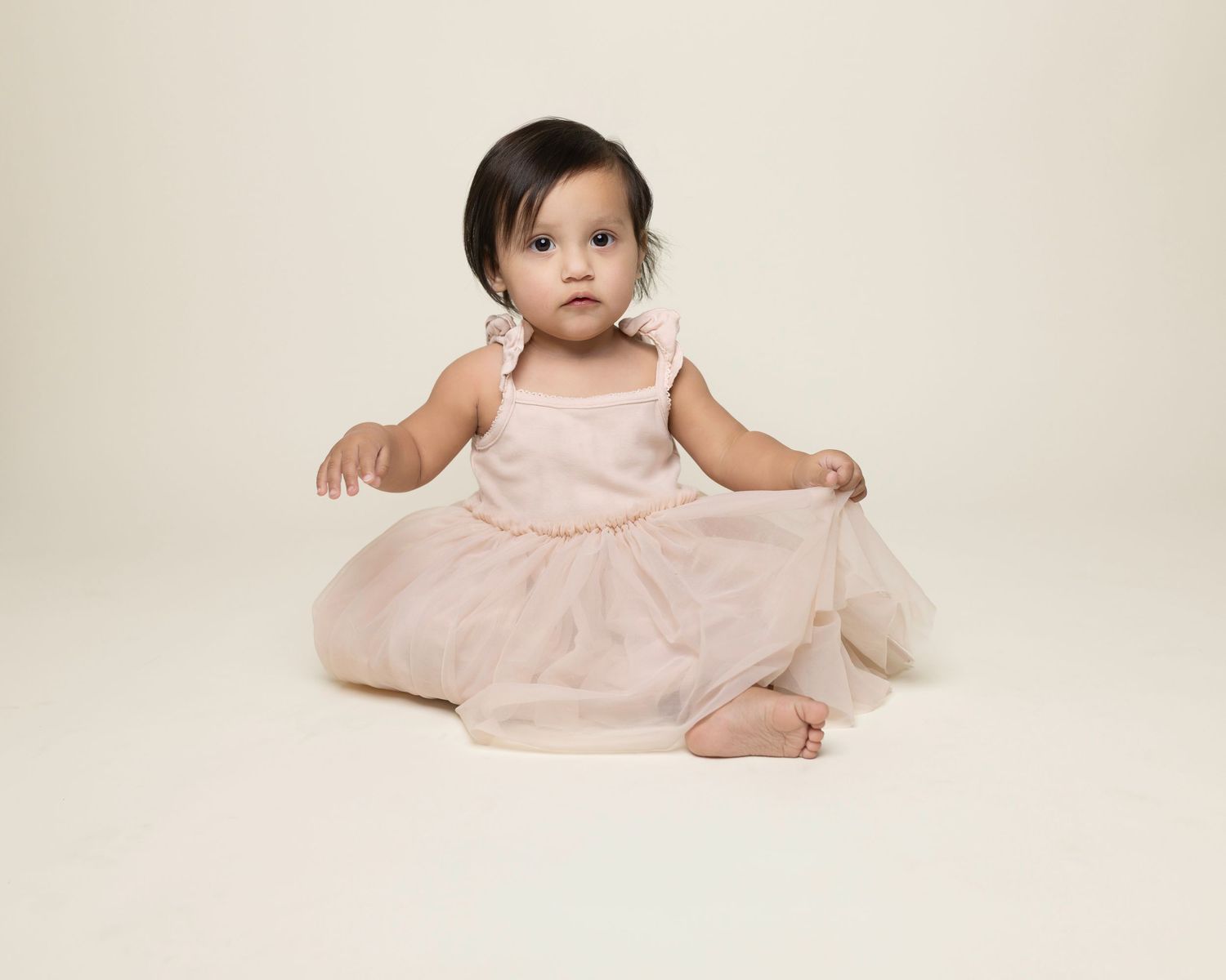 Baby in soft pink dress sits on cream backdrop during professional studio portrait session.