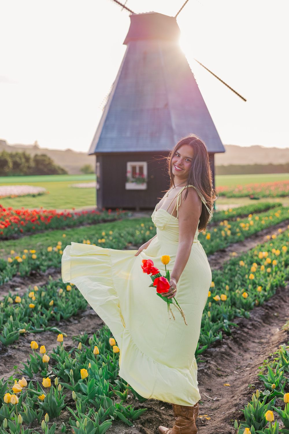 A flowing yellow dress twirls among vibrant tulip fields at sunset with a rustic windmill in the background.
