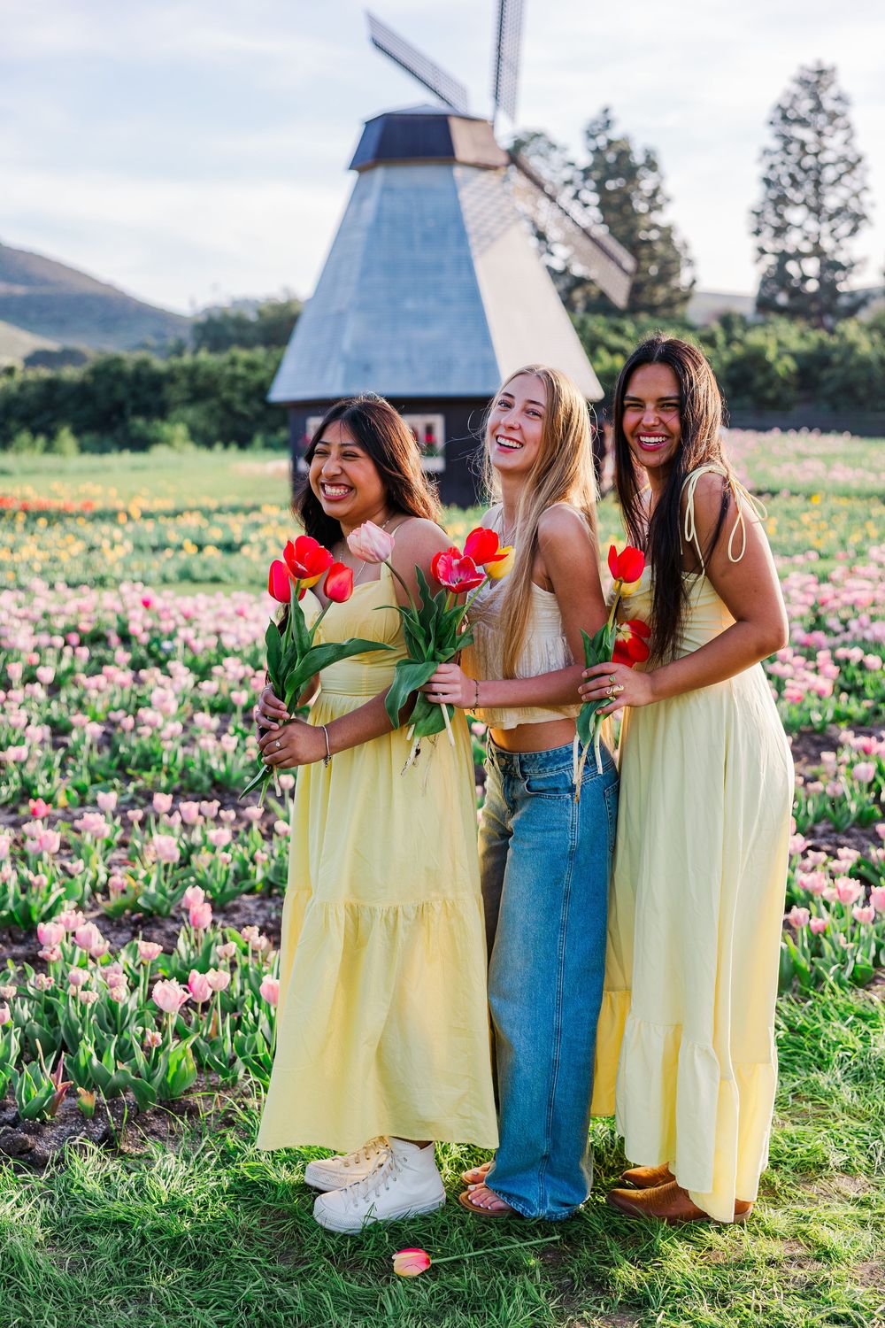 Three friends in yellow dresses and denim stand together laughing in a blooming tulip field on a sunny spring day.