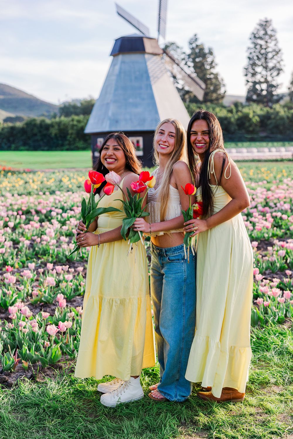 Three friends in yellow dresses and denim enjoy a sunny day in a blooming tulip field next to a windmill.