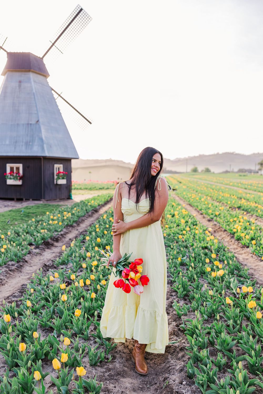 A yellow dress flows in the breeze among vibrant tulip fields with traditional Dutch windmills in the background.