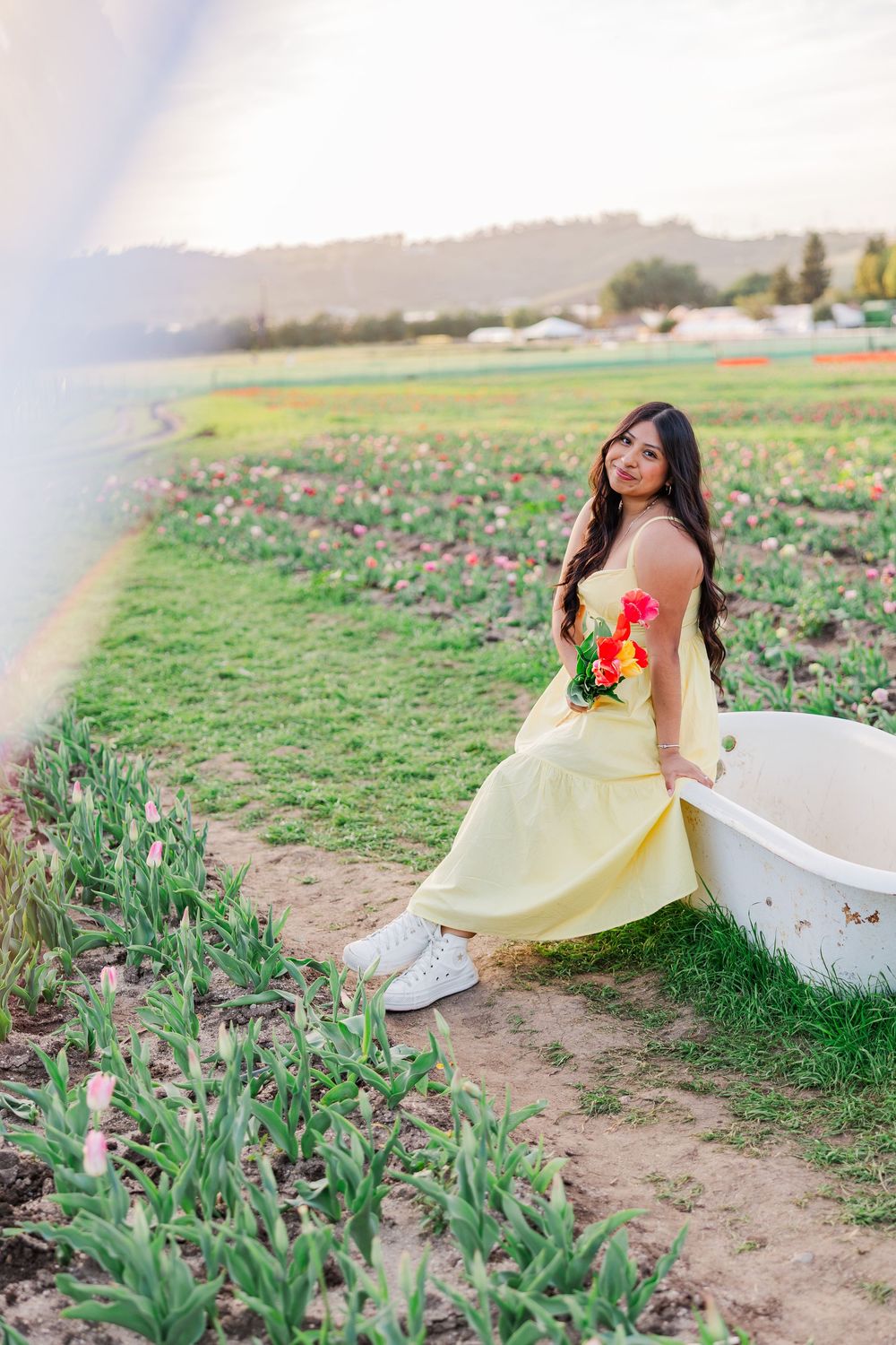 A yellow sundress flows in the breeze as someone poses in a field of spring tulips and flowers.