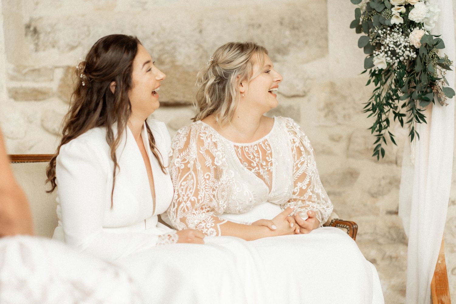 Two people share joyful laughter while sitting together at a rustic stone venue with greenery decorations.