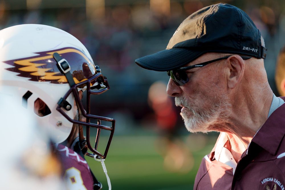 Football coach in a black cap and sunglasses talks closely with a player wearing a maroon and gold helmet.