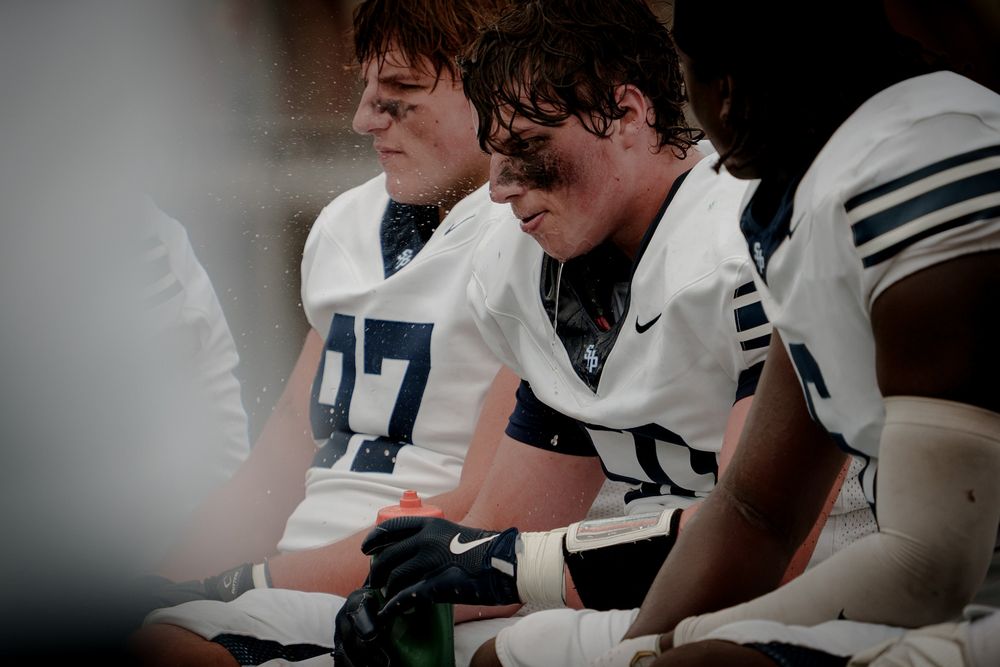 Two football players in white uniforms sit on the sideline bench during a game, appearing focused and tired.