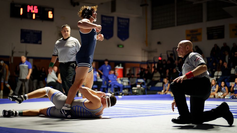 Wrestler celebrates victory as referee raises his hand while opponent lies on mat during a high school wrestling match.