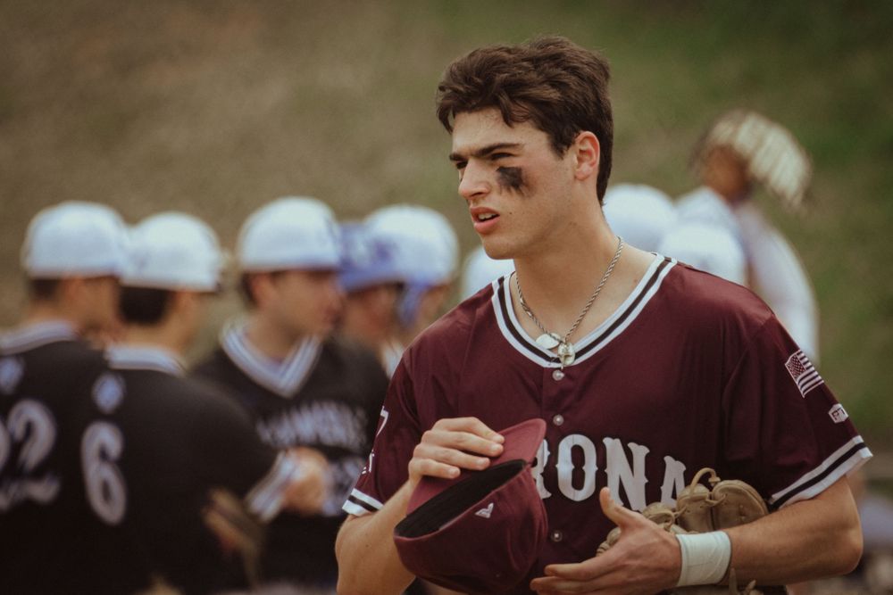 A young baseball player in a maroon Iona uniform holds his helmet during a game.