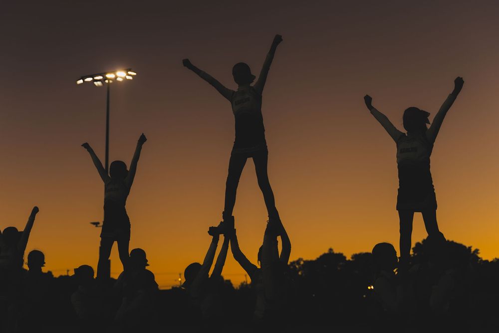 Silhouettes of cheerleaders performing stunts against a golden sunset sky at a sports event.