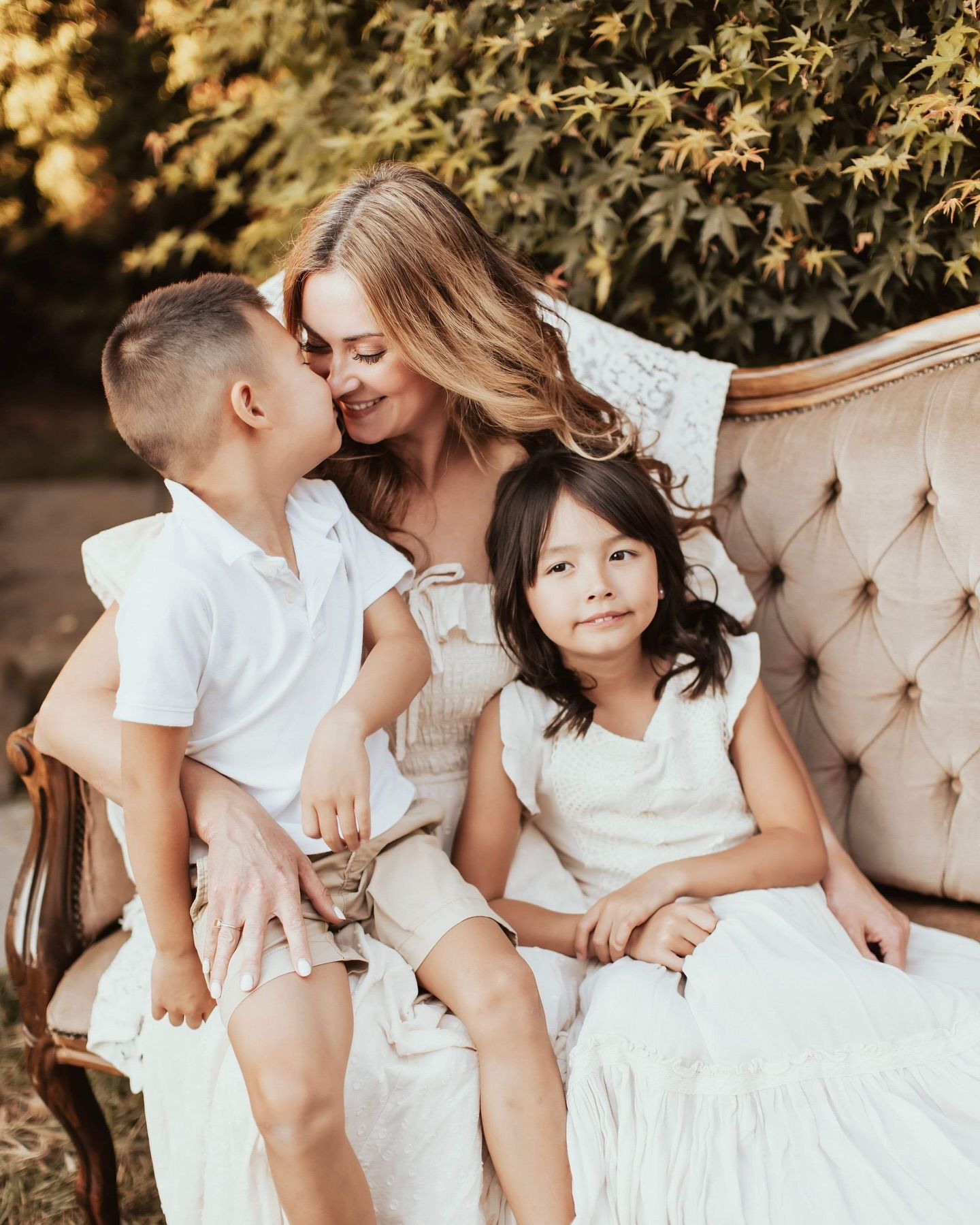A family wearing white sits together on a beige tufted sofa outdoors with autumn foliage in the background.
