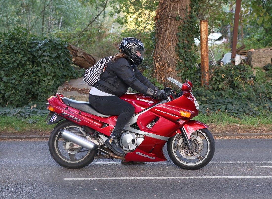A motorcyclist in black riding gear sits on a red sport bike parked on a tree-lined street.