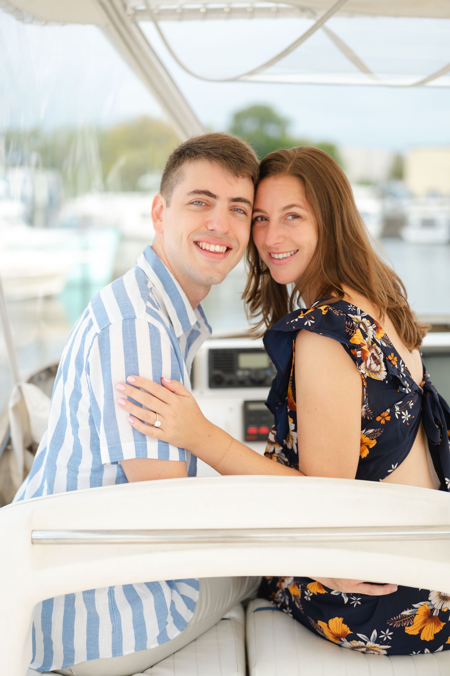 A couple shares a romantic moment together on a boat dock wearing coordinating navy and floral outfits, featuring her new engagement ring.