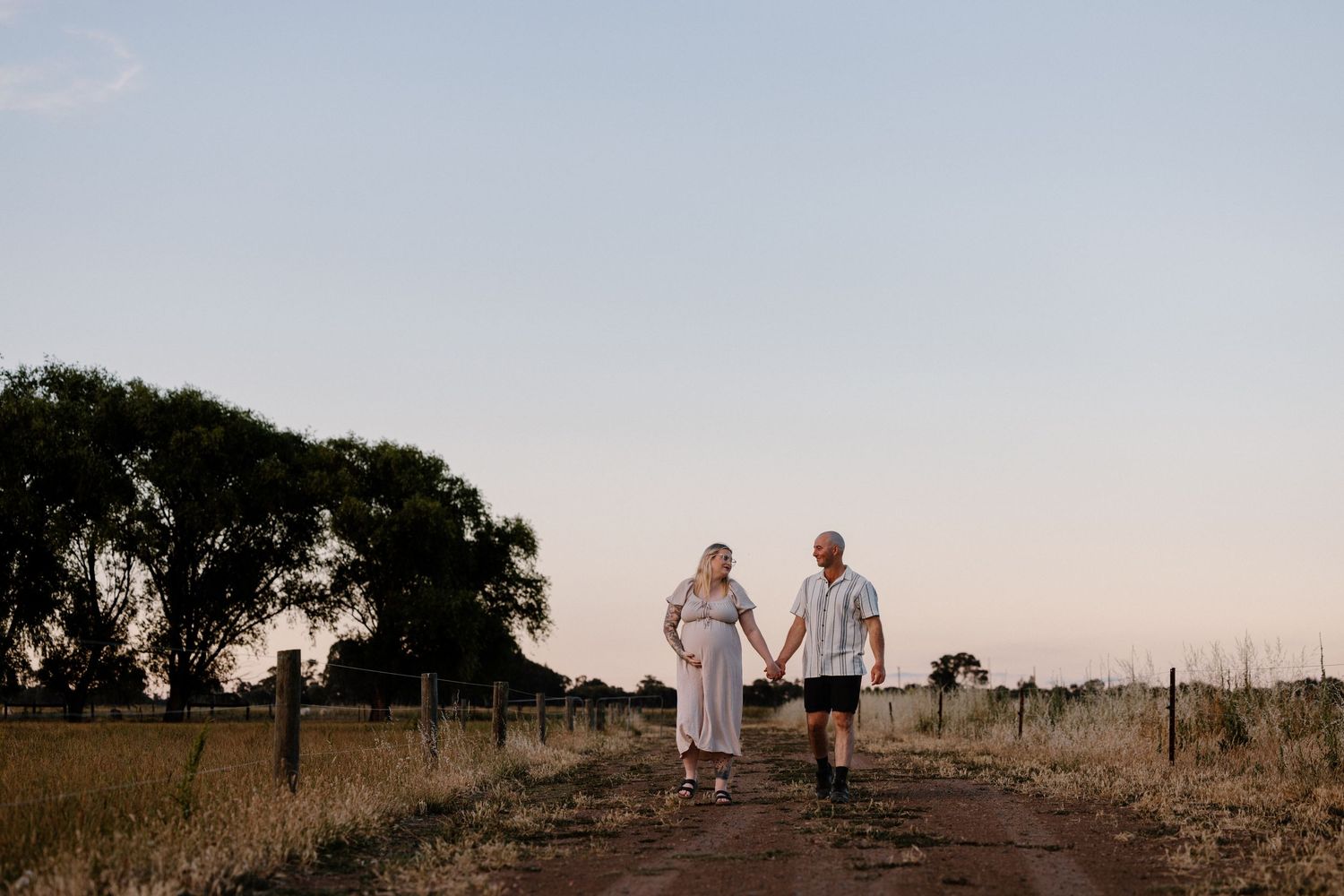 A couple walks hand in hand down a dirt path at sunset with silhouetted trees in the background. location shepparton