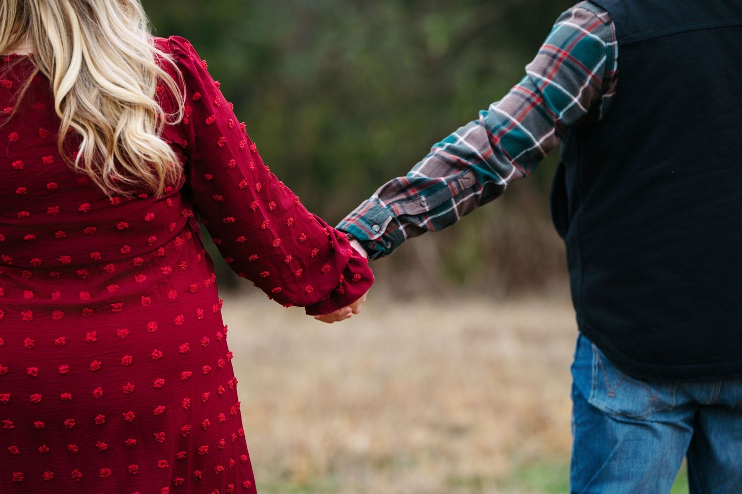 Two people holding hands while wearing a red knit sweater and plaid shirt in an outdoor setting.