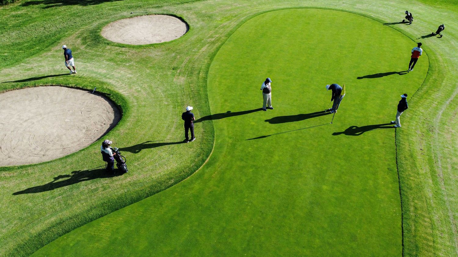 Aerial view of golfers playing on a lush green golf course with sand bunkers and casting long shadows.