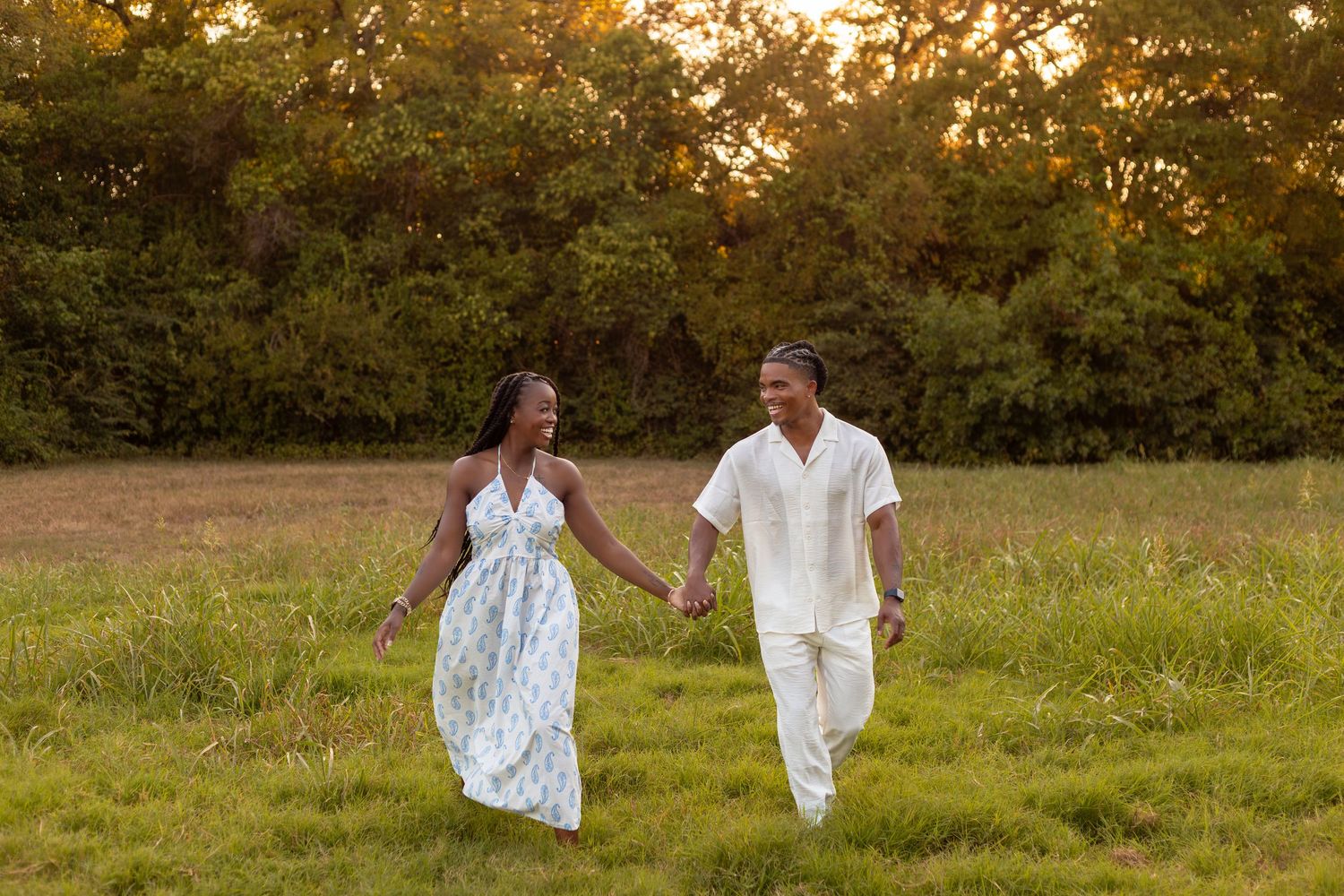 A couple walks hand in hand through a golden sunlit meadow at sunset.