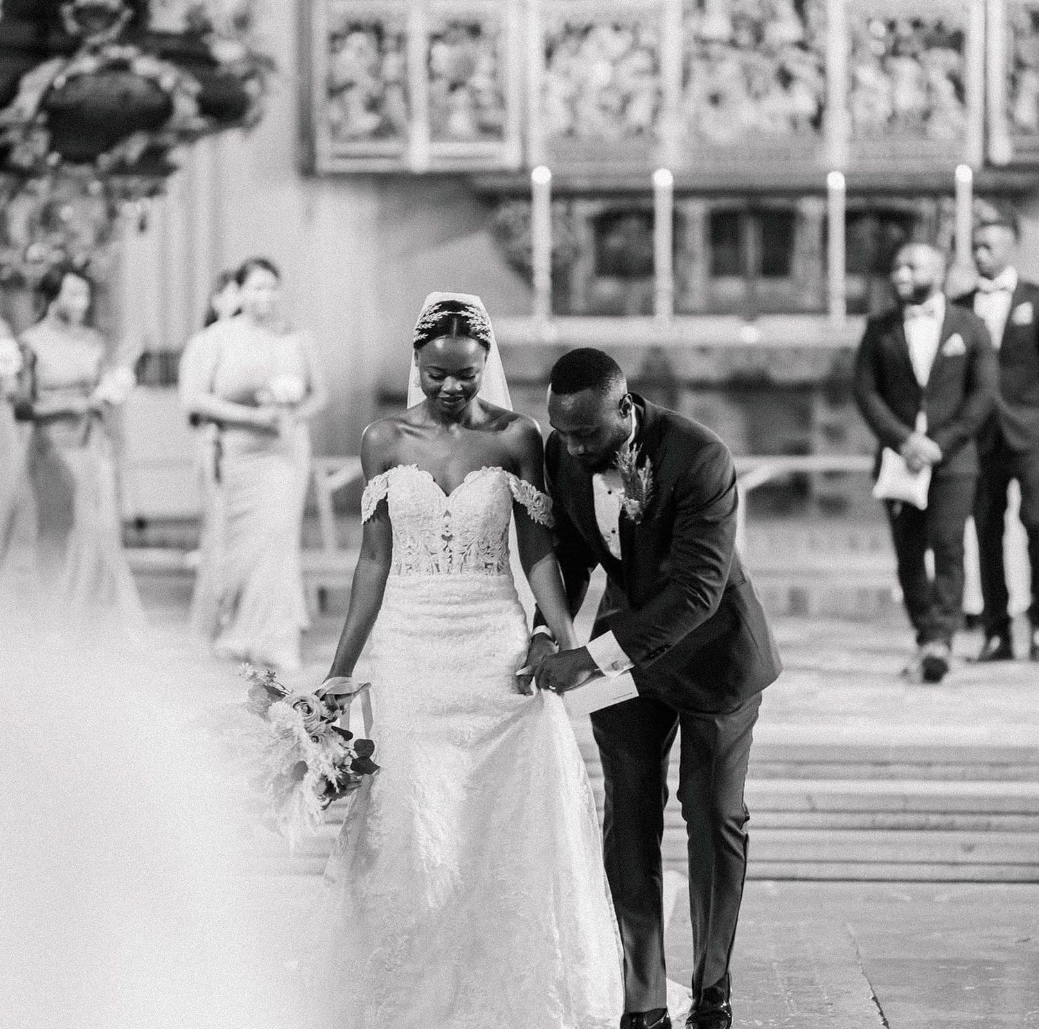 A bride in a flowing white dress and groom in a black suit walk hand in hand along a lakeside path in black and white.