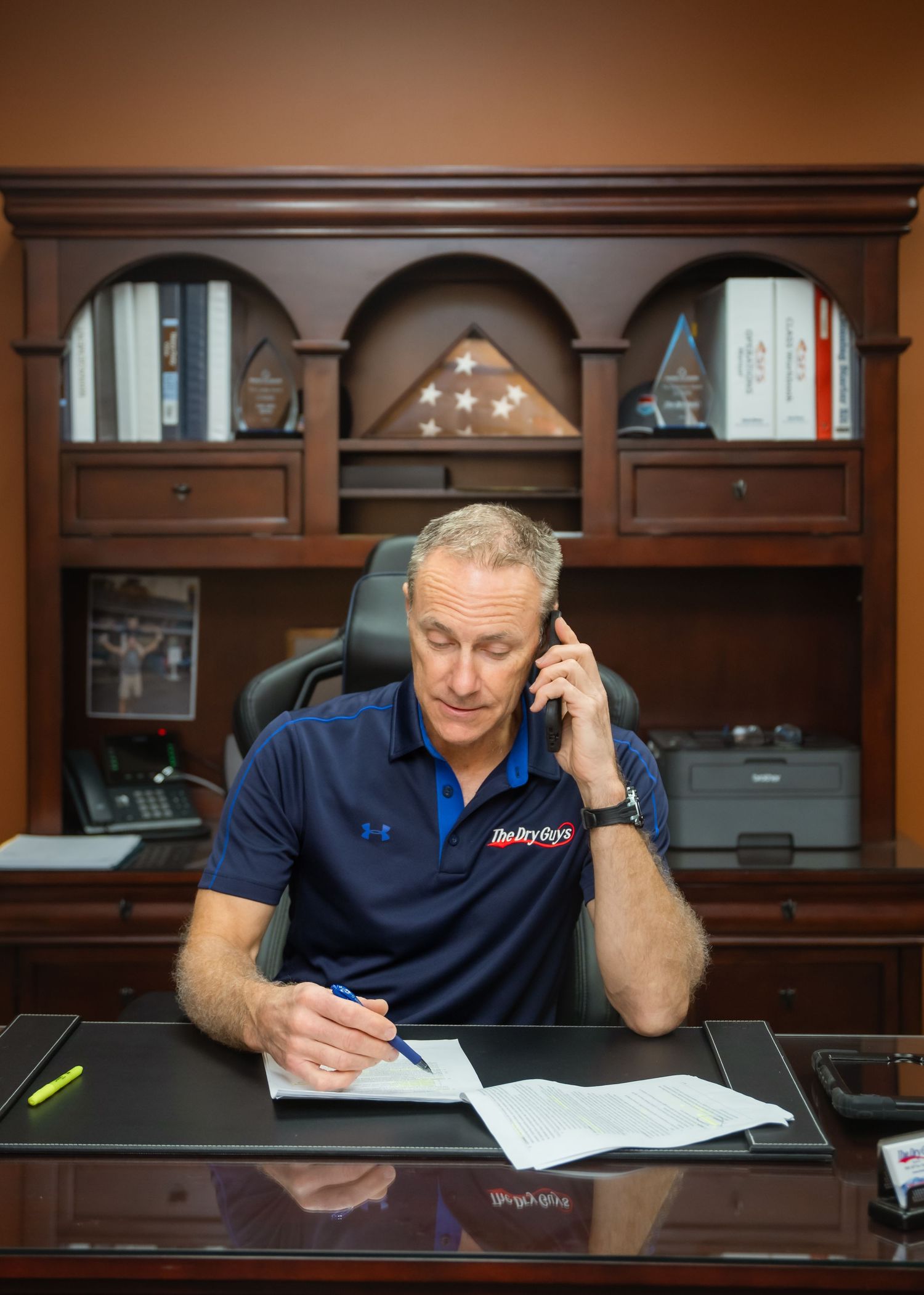 Person in a navy blue shirt sits at a desk making phone calls and reviewing paperwork in an office setting.