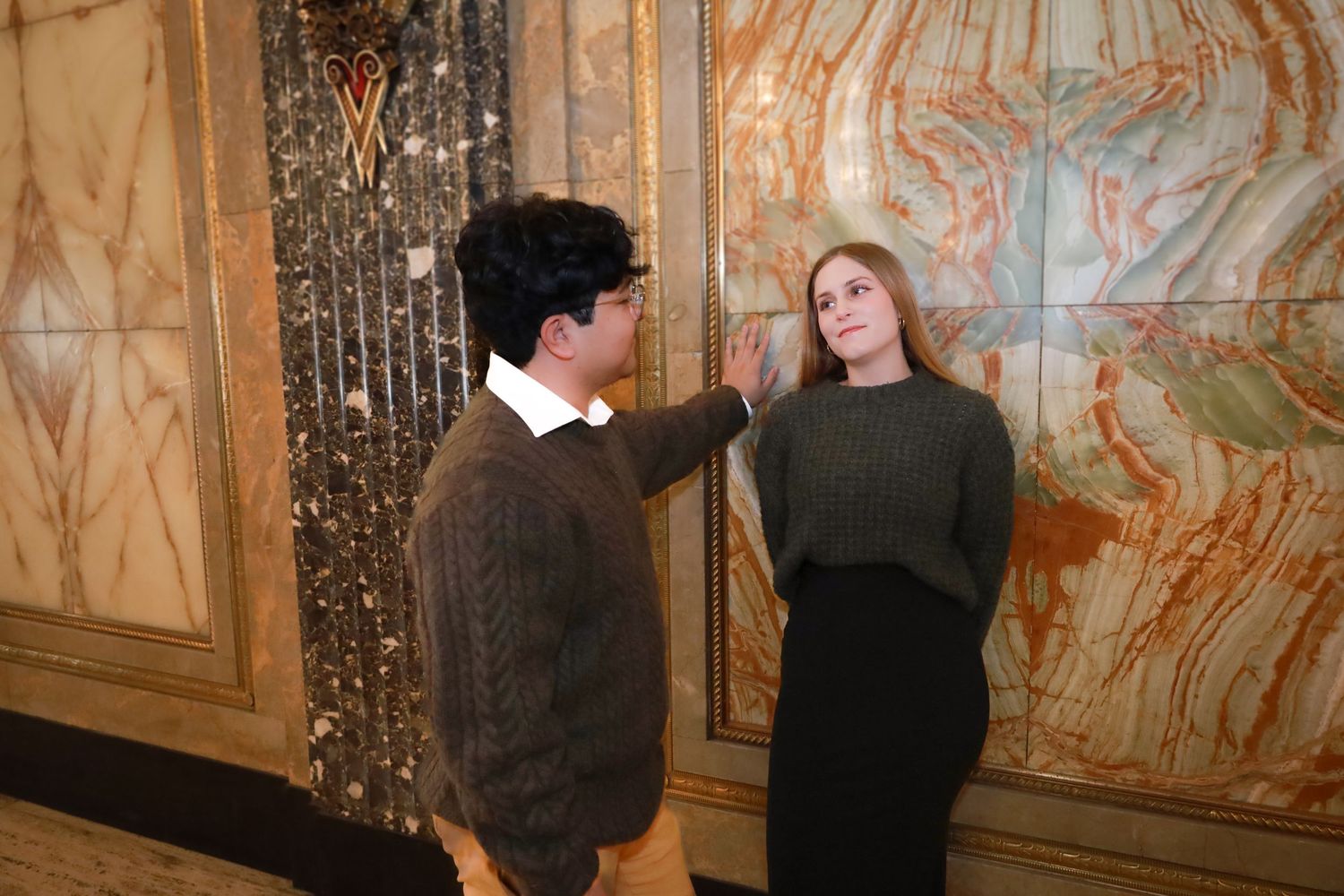 A couple in dark sweaters poses against an ornately decorated wall with gold details.