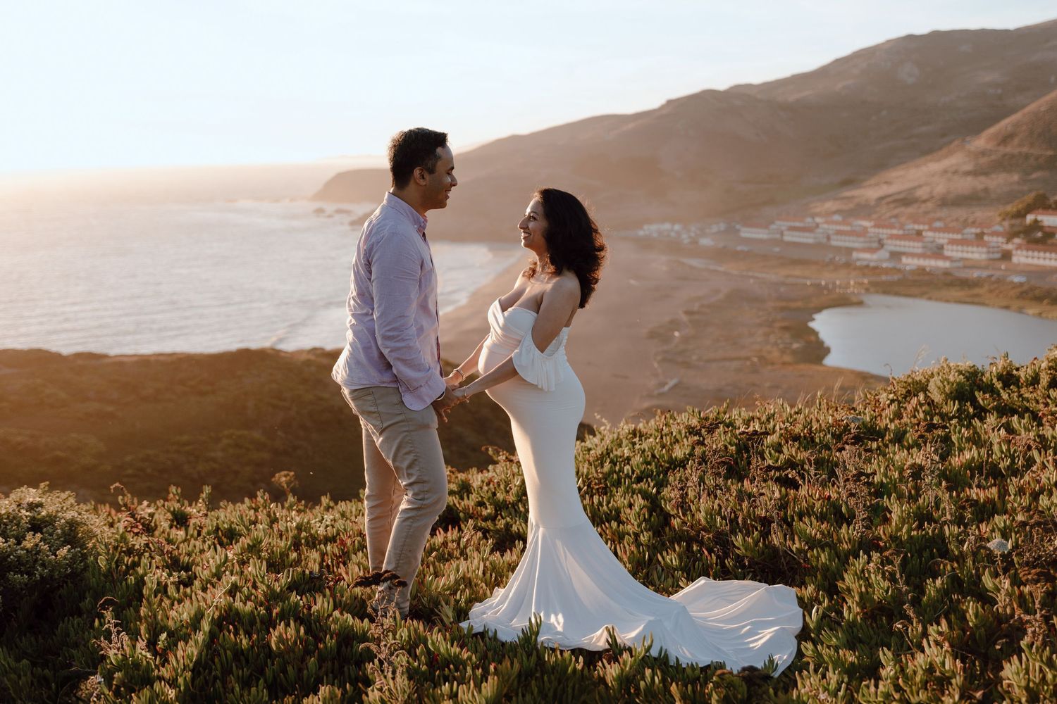 A couple in white attire poses together against a scenic coastal mountain landscape at sunset.