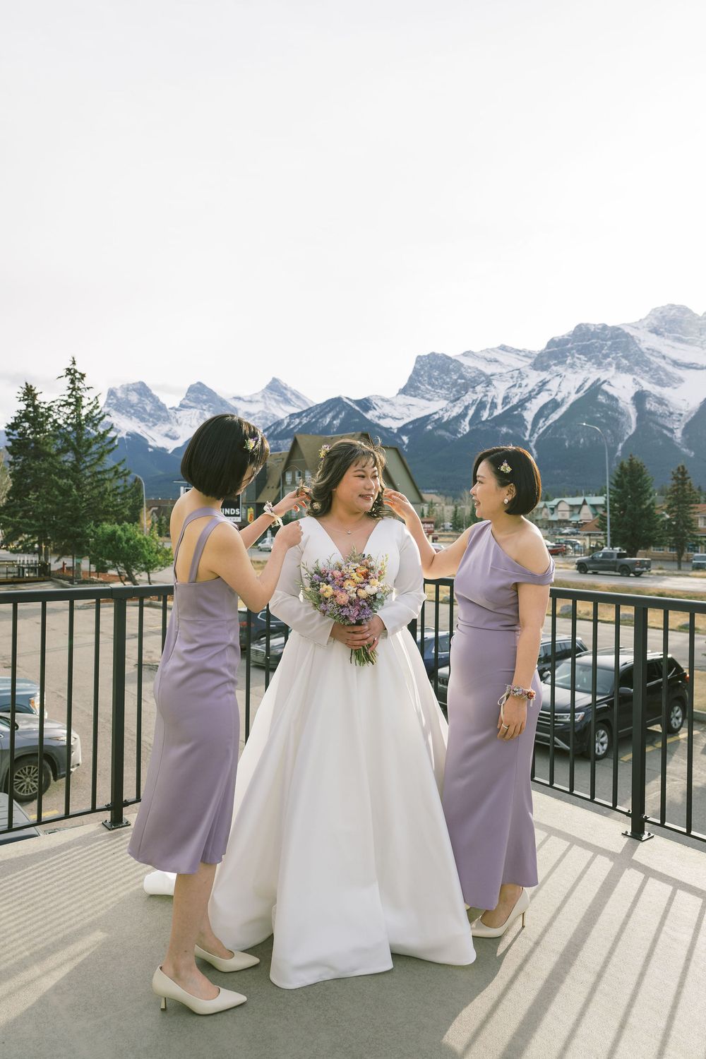 Bridesmaids in lavender dresses and bride in white gown share joyful moments on a balcony with mountain views.