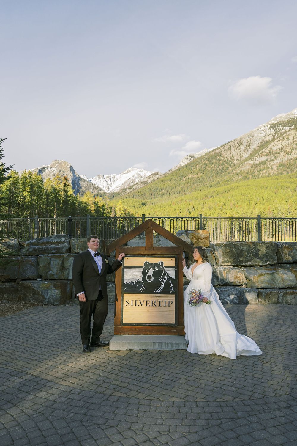 Wedding couple poses with a framed sign against stunning mountain backdrop at outdoor venue with stone walls and patio.