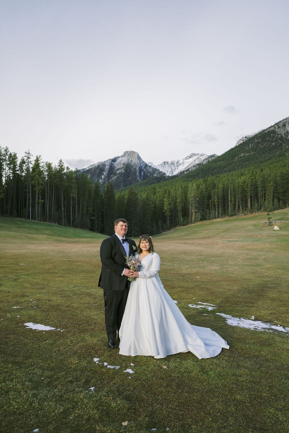 A couple in wedding attire stands in a meadow with snow-capped mountains and evergreen forest in the background.
