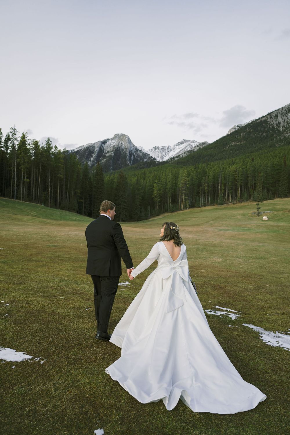 A couple in wedding attire walks across a mountain meadow with snow patches and evergreen forest in the background.