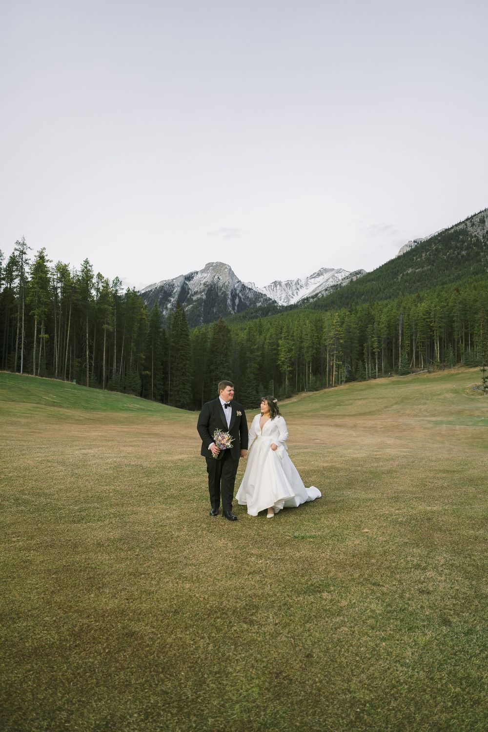 A wedding party walks across a scenic mountain meadow with evergreen forests and snow-capped peaks in the background.