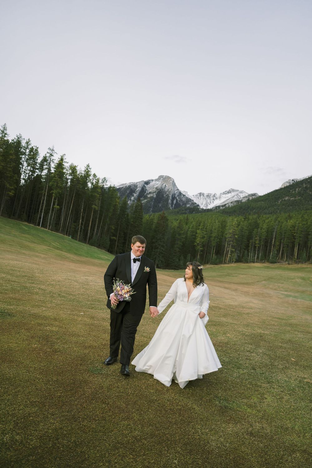 A couple in wedding attire walks across a scenic mountain meadow with evergreen forest and snow-capped peaks in the background.