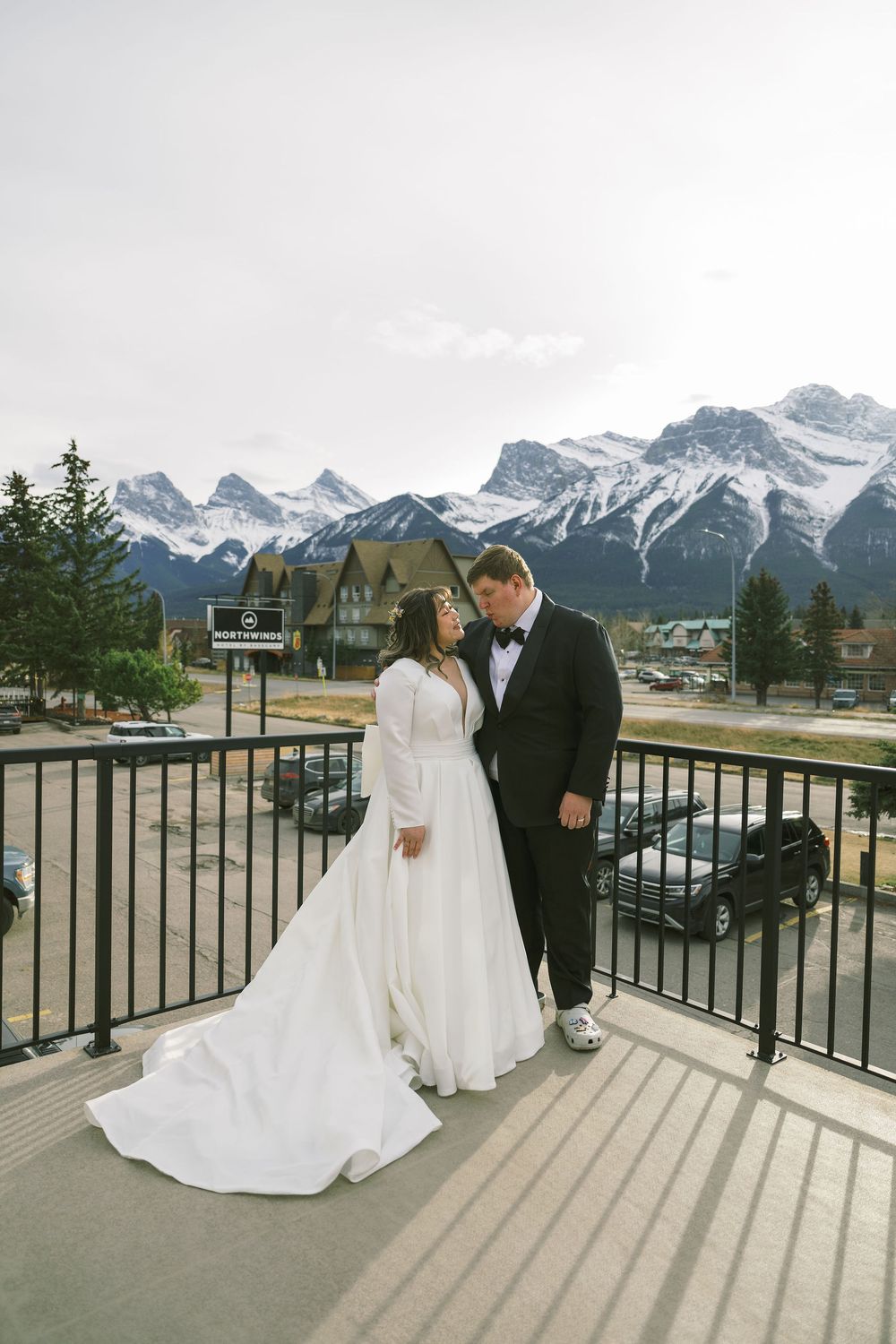 A couple in wedding attire stands on a deck overlooking snow-capped mountain peaks in the Canadian Rockies.