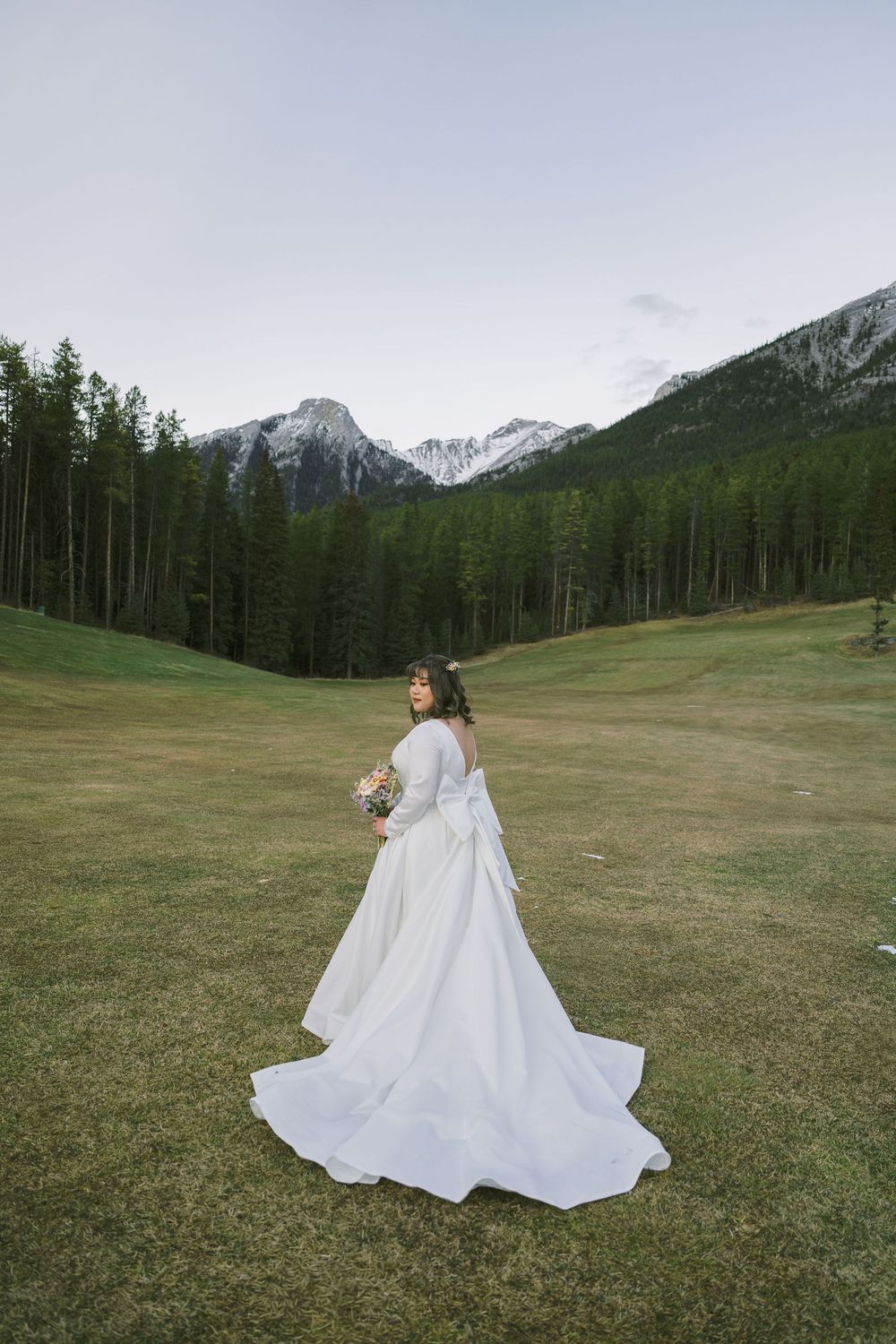 A figure in a flowing white wedding dress twirls in a mountain meadow with snow-capped peaks and evergreen forests behind.