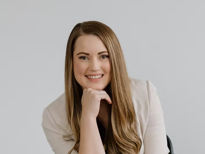 Professional headshot of someone in a light colored blazer with long brown hair against a neutral background.
