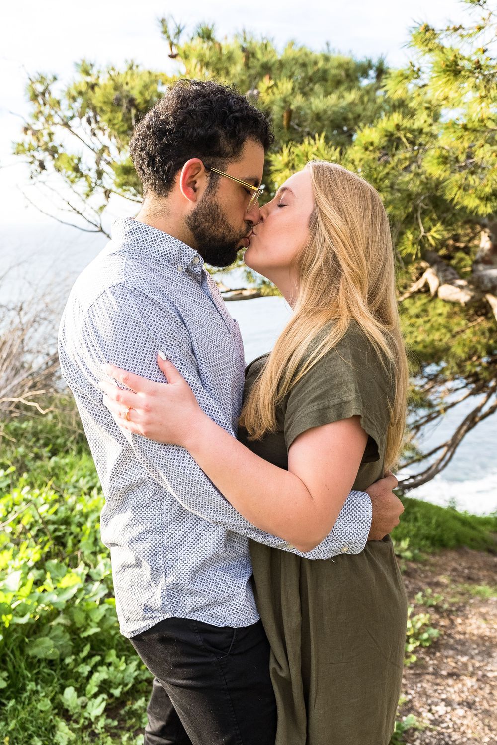A romantic couple shares a tender kiss outdoors against a backdrop of green foliage and natural scenery.