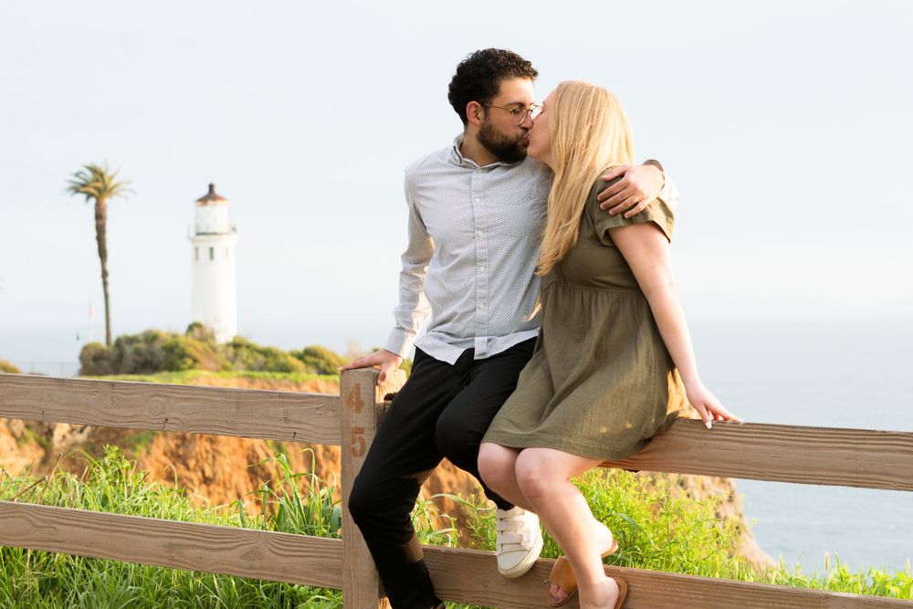 A couple sits together on a wooden railing with a lighthouse and ocean cliffs in the background.