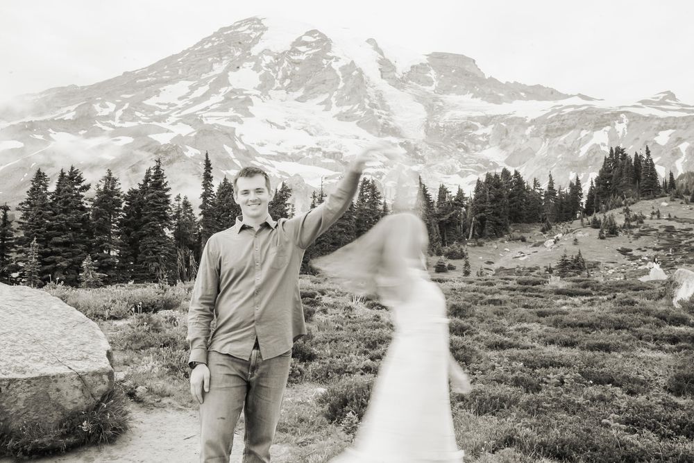 A couple walks along a winding mountain trail with Mount Rainier's majestic peak in the distance.