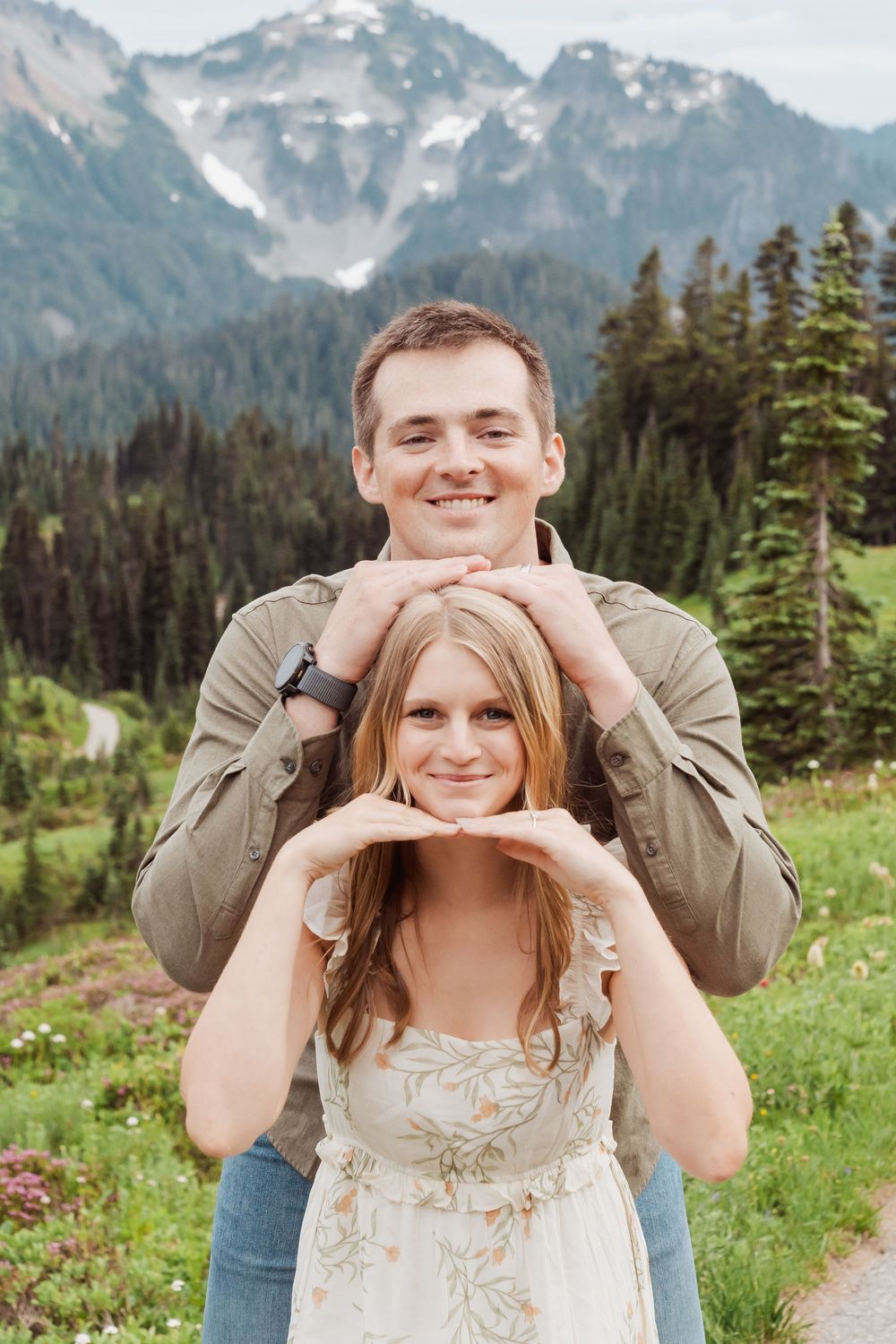 A couple poses playfully in a meadow with snow-capped mountains and evergreen forests creating a scenic backdrop.