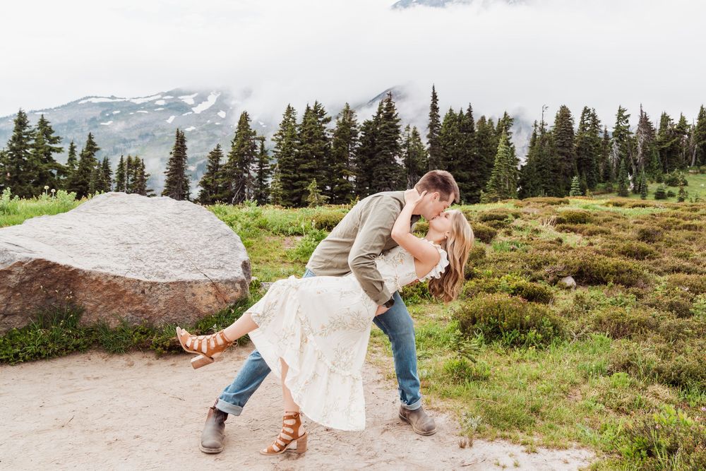 A scenic mountain trail leads through alpine meadows with snow-capped peaks and evergreen trees in the background.