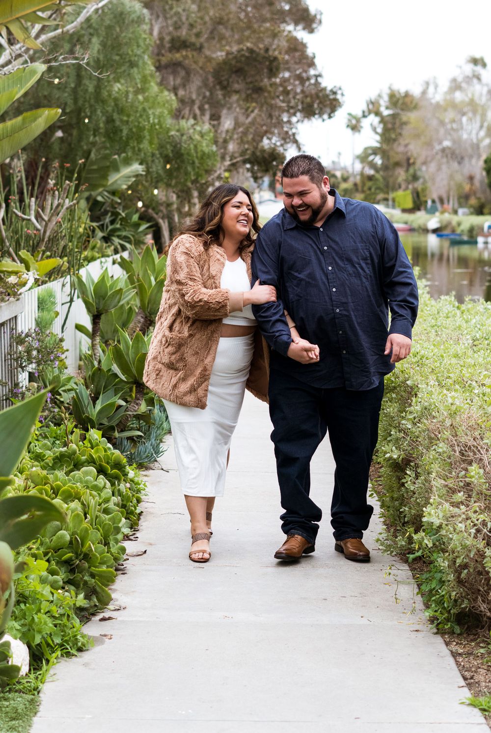 A couple walks together on a garden path surrounded by lush greenery on a sunny afternoon.