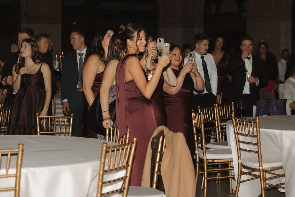 Wedding guests in burgundy dresses and formal attire gather around tables with gold chiavari chairs at an elegant reception.