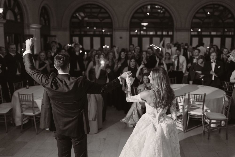 Elegant ballroom wedding reception with guests seated at round tables under arched windows.