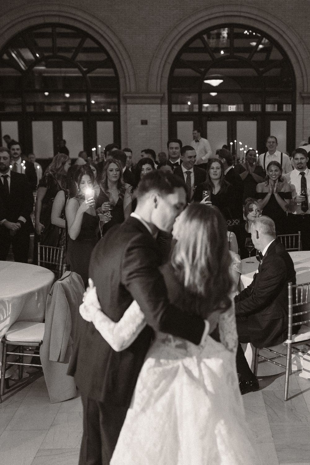 Wedding guests dance and celebrate at an elegant indoor reception with arched windows.