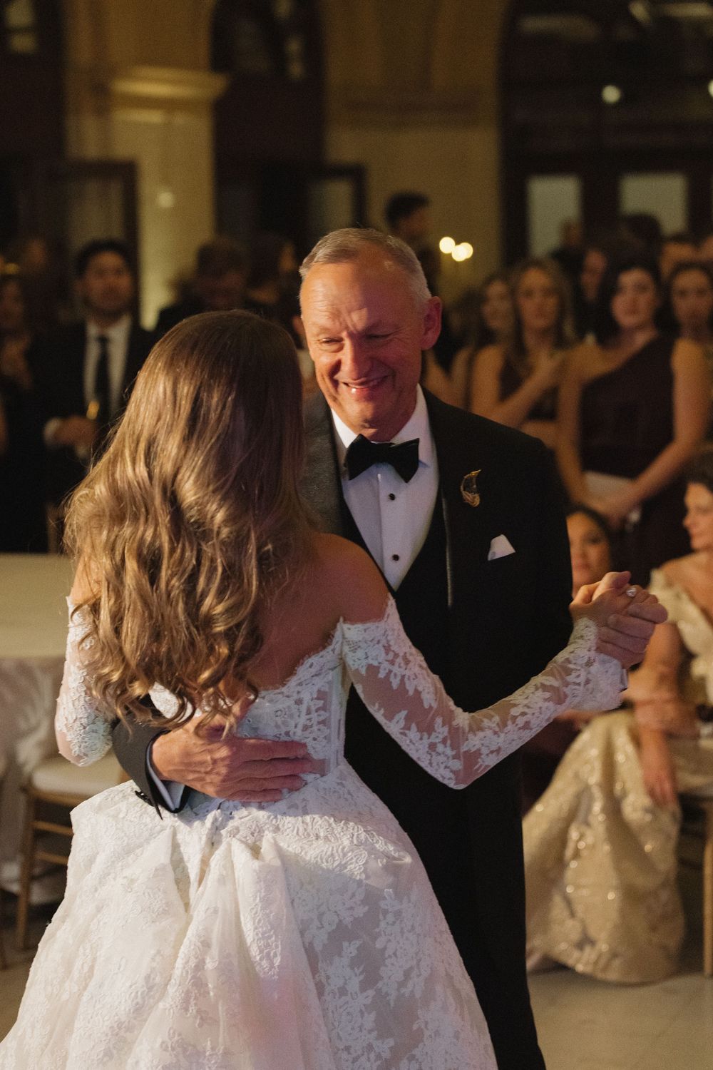 Wedding couple performs their first dance in a beautifully lit ballroom surrounded by guests.