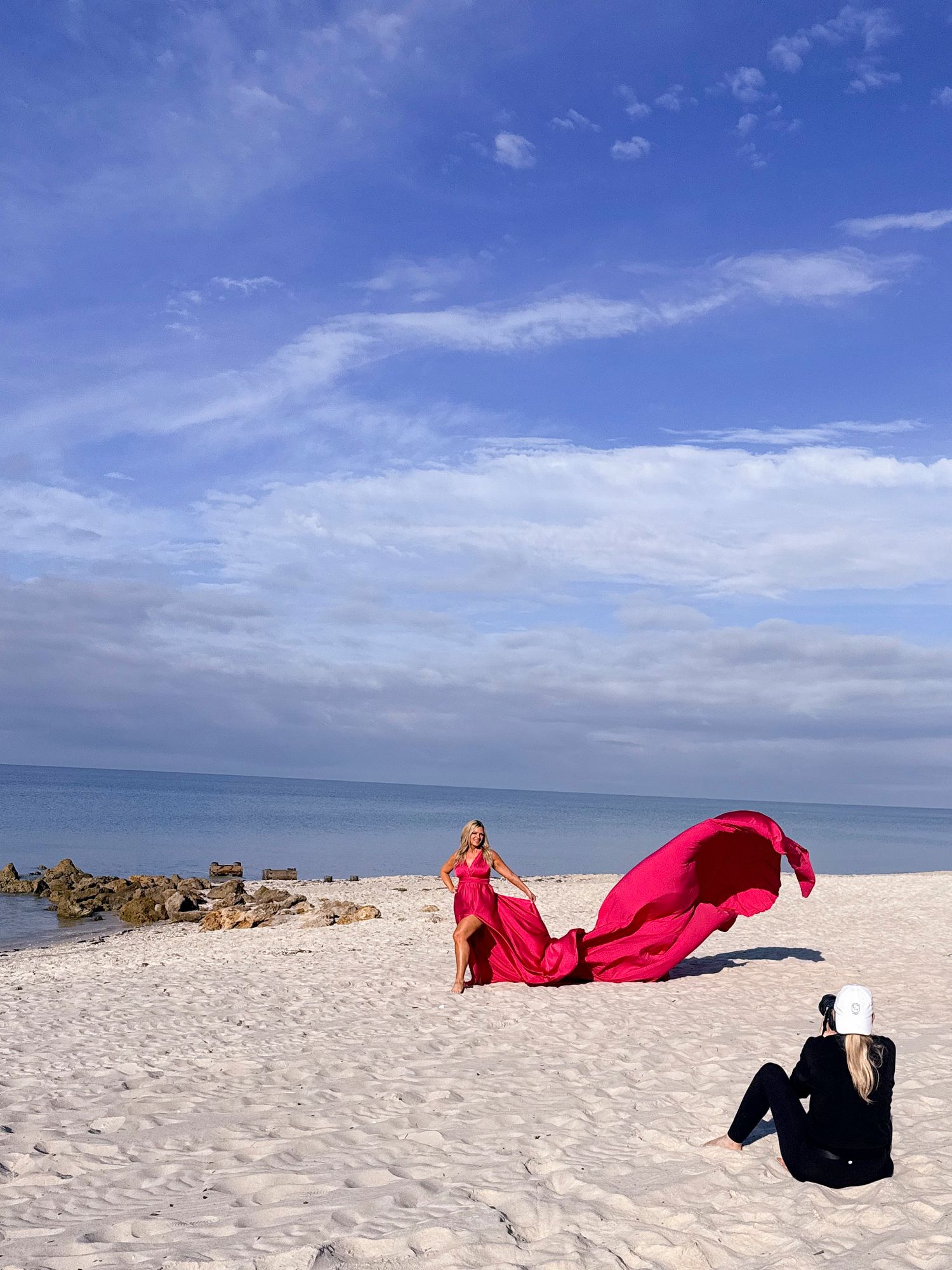 A flowing red dress billows in the wind on a pristine white sand beach against a bright blue sky and ocean backdrop.
