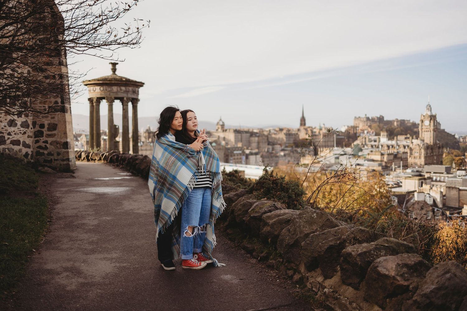 Two people sit on stone wall overlooking historic Edinburgh cityscape during golden hour.
